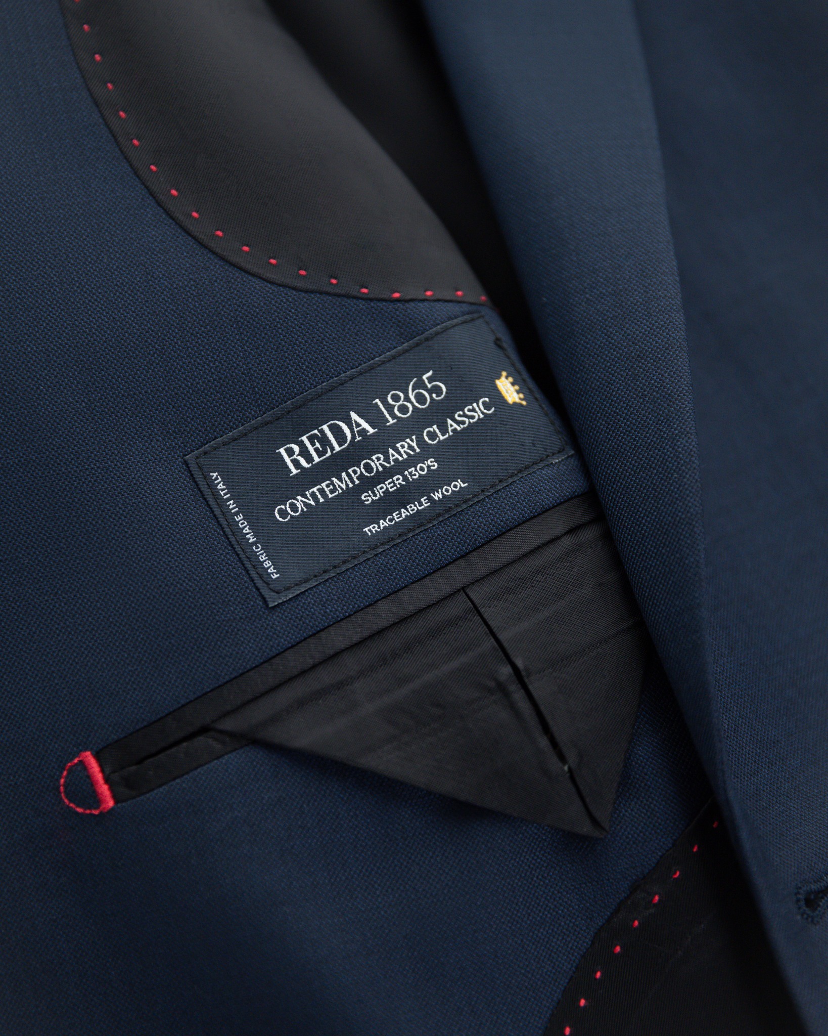 西裝內襯局部特寫，展示黑色織標印有 REDA 1865 CONTEMPORARY CLASSIC Super 130's TRACEABLE WOOL 字樣，周邊延伸著密集的紅色手針點腳縫線，灰色裡布折疊出挺拔的三角袋蓋細節